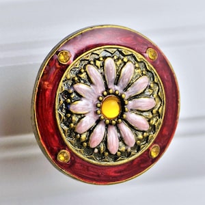 May include: Decorative cabinet knob with a floral design. The knob features a central daisy with pink petals and a yellow center, surrounded by ornate gold detailing. The outer ring is a deep red with small yellow accents.