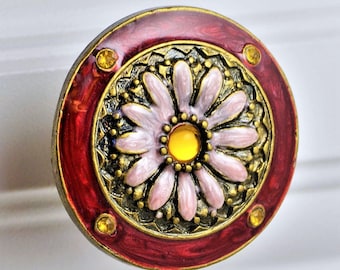 May include: Decorative cabinet knob with a floral design. The knob features a central daisy with pink petals and a yellow center, surrounded by ornate gold detailing. The outer ring is a deep red with small yellow accents.
