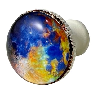 May include: A round, decorative cabinet knob with a celestial design. The knob features a detailed image of a planet with blue, yellow, and white hues, set within a silver-colored, textured bezel. The knob has a white, spherical base.