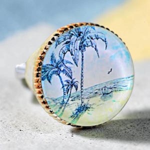 May include: A round ring featuring a gold-toned metal band with a textured edge. The face of the ring displays a circular illustration of a beach scene with blue palm trees, a sandy shore, a calm ocean, and a small bird flying in the sky.