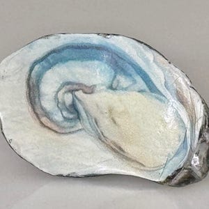 May include: An oyster shell painted with watercolor. The shell features a creamy white interior with blue and gray swirls. The outer edge is a dark gray color. The artwork is a close-up view of the shell.