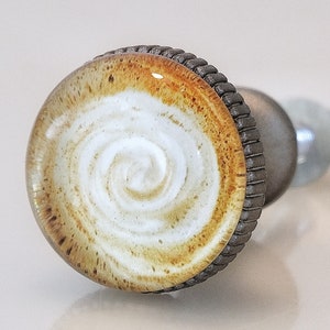 May include: A decorative cabinet knob with a coffee-inspired design. The knob features a circular glass top with a swirl of white and brown, resembling a latte. The knob has a dark metal base and a textured edge.