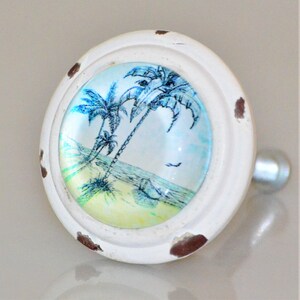May include: White, distressed cabinet knob with a beach scene. The knob features a glass insert with a blue and green tropical beach design, including palm trees, sand, and ocean. The knob has a silver-colored handle.