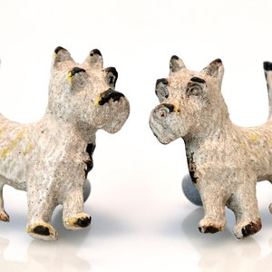 May include: A pair of dog-shaped cufflinks. The cufflinks are off-white with black and yellow accents. The dogs are standing, with their tails up. The cufflinks have a textured appearance and are set on a white background.