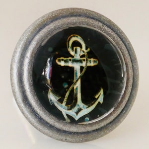 May include: A round, silver-toned cabinet knob with a nautical theme. The knob features a clear, domed center with a teal and white anchor design set against a black background. The knob has a textured, metallic appearance.