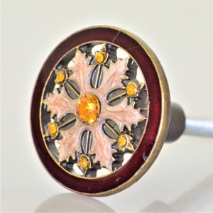 May include: Decorative cabinet knob with a circular design. The knob features a dark red outer ring, a gold-colored metal frame, and a central design with light pink and black accents. It is adorned with yellow-orange gemstones.