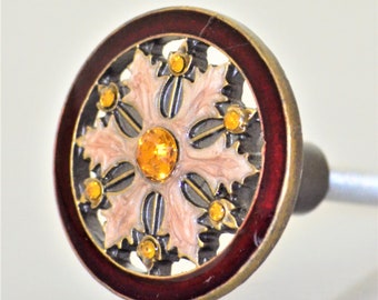 May include: Decorative cabinet knob with a circular design. The knob features a dark red outer ring, a gold-colored metal frame, and a central design with light pink and black accents. It is adorned with yellow-orange gemstones.