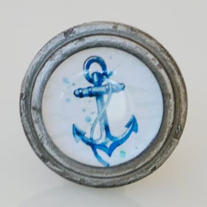 May include: A round, nautical-themed cabinet knob with a distressed silver finish. The center features a blue anchor design on a white background, with blue watercolor-style accents. The knob is likely made of metal and glass.