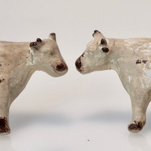 May include: Two small, off-white cow figurines with dark brown spots and accents. The cows are standing side-by-side, facing each other. The figurines have a rustic, aged appearance, suggesting they are handcrafted or vintage decorative items.