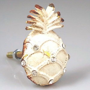 May include: A decorative pineapple-shaped cabinet knob with a distressed, off-white finish. The knob features a textured surface, brown accents, and is embellished with clear, sparkling rhinestones. The hardware includes a metal bolt and nut.