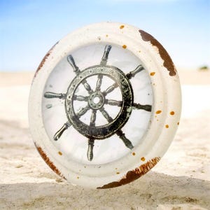 May include: A round decorative knob featuring a central illustration of a dark gray ship's wheel. The knob has a distressed off-white finish with brown paint splatters and worn edges, creating a textured appearance around the circular center.