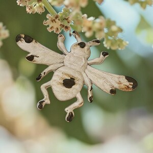 May include: A wooden insect ornament featuring a light beige finish with dark brown painted spots on the wings, head, and body. The insect has six legs, two antennae, and four wings, with a distressed texture throughout the surface of the wood.