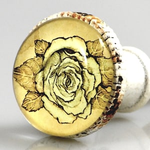 May include: A decorative cabinet knob featuring a rose design. The knob has a clear, rounded glass top with a yellow rose illustration and a light yellow background. The edges are textured with a brown and white pattern.