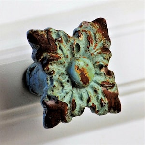 May include: A decorative, antique-style cabinet knob with a floral design. The knob features a distressed finish with a turquoise base and brown accents, giving it a rustic appearance. The knob is mounted on a white surface.