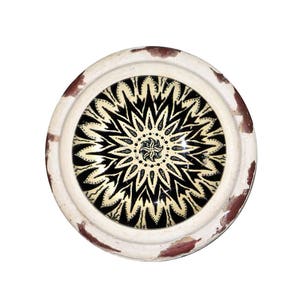 May include: Round decorative plate with a black and cream floral design in the center. The plate has a white rim with distressed brown accents. The design is symmetrical and detailed, creating a striking visual.