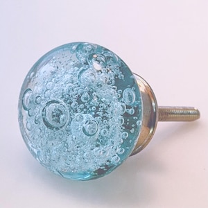 May include: A round cabinet knob featuring a clear, light blue glass sphere filled with numerous small, trapped air bubbles. The glass sphere is attached to a metallic base with a threaded screw extending from the back for installation.