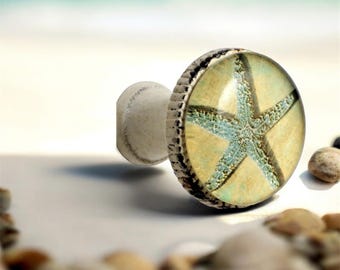 May include: A round cabinet knob featuring a light blue starfish graphic set against a beige background. The knob has a metallic, textured silver-toned rim and a smooth, rounded white base, providing a functional handle for furniture drawers.