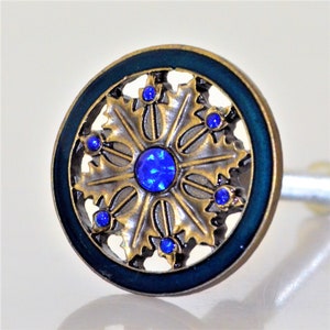 May include: A decorative cabinet knob with a circular design. The knob features a dark teal border, a gold-colored filigree pattern, and is adorned with blue gemstones. The center and surrounding accents are a vibrant blue.