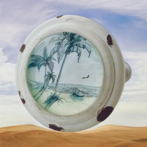 May include: A round ceramic knob featuring a hand-painted scene of palm trees on a beach with a light blue sky. The white border of the knob is decorated with several dark brown, irregular spots, giving it a weathered and rustic appearance.
