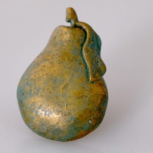 May include: A pear-shaped decorative object with a textured surface. The pear is primarily gold with patches of teal-colored accents. A stem and leaf are attached to the top of the pear.