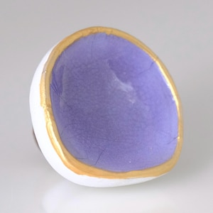 May include: A handcrafted ceramic ring with a white exterior, a lavender interior, and a gold-colored rim. The ring has an organic, irregular shape, and is likely a piece of jewelry or a decorative item.