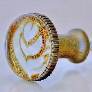 May include: A decorative cabinet knob with a coffee art design. The knob features a clear, rounded top with a brown and white latte art pattern. The base and stem are a bronze color, and the edge of the knob has a textured, ridged design.