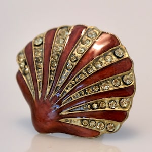 May include: A decorative seashell-shaped object with a reddish-brown enamel finish and a gold-colored metal trim. The shell is adorned with rows of clear, sparkling rhinestones, adding a touch of glamour to the design.