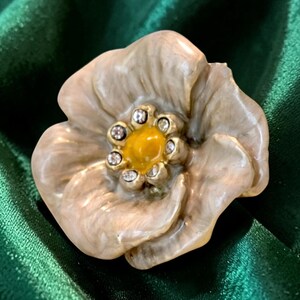 May include: A flower-shaped brooch featuring light beige, swirled enamel petals with a glossy finish. The center of the flower is a smooth, bright yellow circular accent surrounded by a ring of small, clear, sparkling rhinestones set in metal.