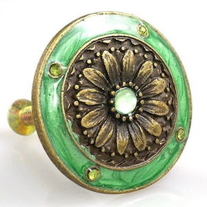 May include: Decorative cabinet knob with a floral design. The knob features a bronze-colored flower with a green enamel border and a central green gem. Small green gems are set around the border. The knob is attached to a gold-colored screw.