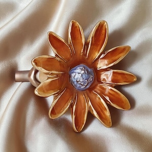 May include: A decorative wall hook featuring a large, glossy, orange-brown flower with elongated petals and a textured, light purple center. The flower is attached to a smooth, cylindrical, metallic-toned mounting base extending to the left side.