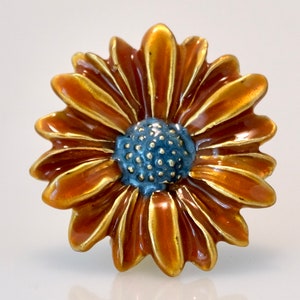 May include: A detailed, close-up view of a flower-shaped brooch. The brooch features a central blue-gray disc surrounded by orange-brown petals with gold-colored edges. The design is highly detailed, with a glossy finish.