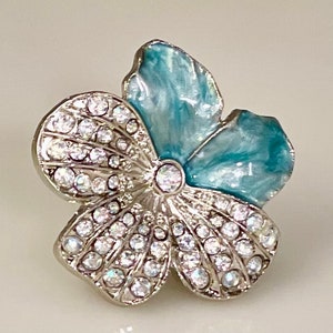 May include: A flower-shaped brooch with a silver-tone metal base. The brooch features clear, sparkling crystals and two petals with a light blue enamel finish. The brooch is a jewelry item.