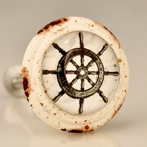 May include: A nautical-themed cabinet knob with a ship's wheel design. The knob features a white, distressed finish with brown accents and a clear glass overlay showcasing the wheel. The hardware is a decorative accent for drawers or cabinets.