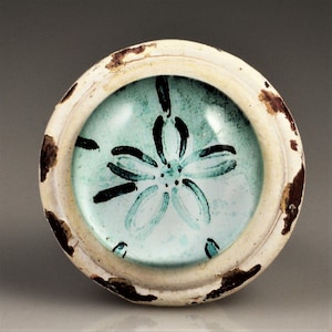May include: Round, off-white ceramic dish with a distressed, aged appearance. The center features a light blue background with a black floral design. The rim has brown spots, adding to the vintage aesthetic.