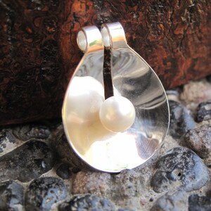 May include: A silver pendant with two white pearls inside. The pendant is shaped like a teardrop and has a smooth, shiny surface.