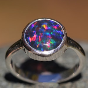Peut inclure: Une bague en argent avec une opale noire ronde sertie dans un sertissage à bélière. L'opale présente des reflets de rouge, de vert et de bleu.