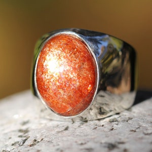 May include: A silver ring with a large, oval-shaped sunstone. The sunstone is a deep orange color with flecks of gold.