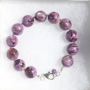 May include: A bracelet made of large, round, purple beads with dark purple and brown swirling patterns. The beads are strung on a purple cord and secured with a silver-coloured clasp. The bracelet is displayed in a white box.