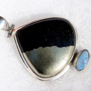 May include: A silver pendant with a black and gold gemstone and a smaller opal gemstone. The pendant is in the shape of a teardrop.