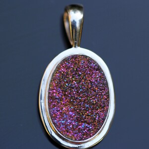 Puede incluir: Un colgante de plata con una piedra preciosa iridiscente de forma ovalada. La piedra preciosa es de color morado oscuro con toques de oro y azul.
