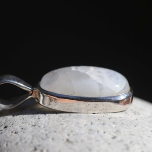 May include: A silver pendant with a white moonstone cabochon. The pendant is oval shaped and has a smooth, polished finish.