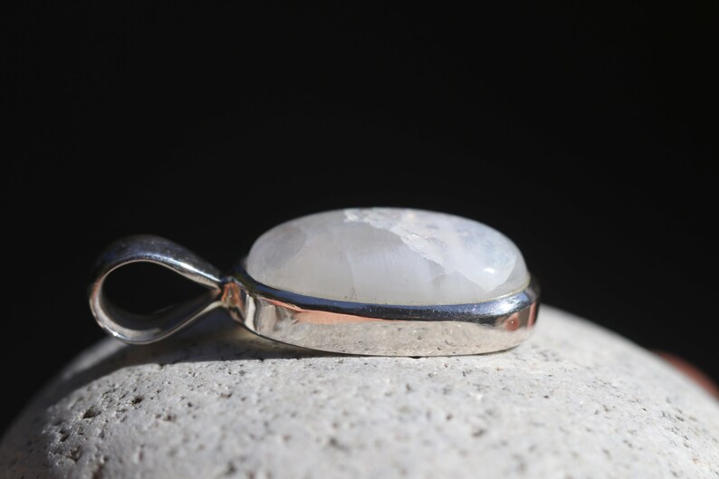 May include: A silver pendant with a white moonstone cabochon. The pendant is oval shaped and has a smooth, polished finish.