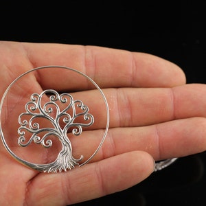 May include: Silver hoop earrings featuring a tree of life design. The tree is crafted from delicate swirls and branches.