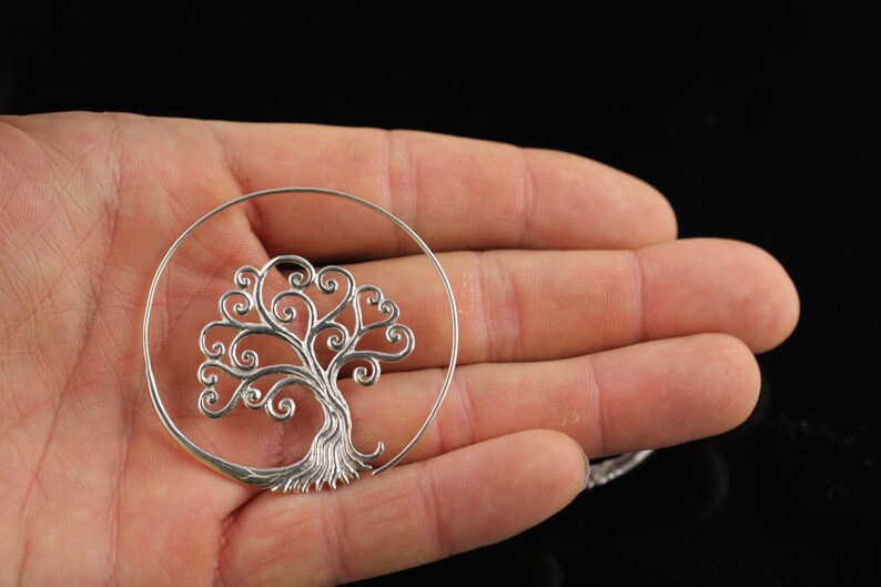 May include: Silver hoop earrings featuring a tree of life design. The tree is crafted from delicate swirls and branches.