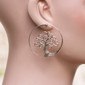 May include: Silver hoop earrings featuring a tree of life design. The tree is crafted from delicate swirls and branches.