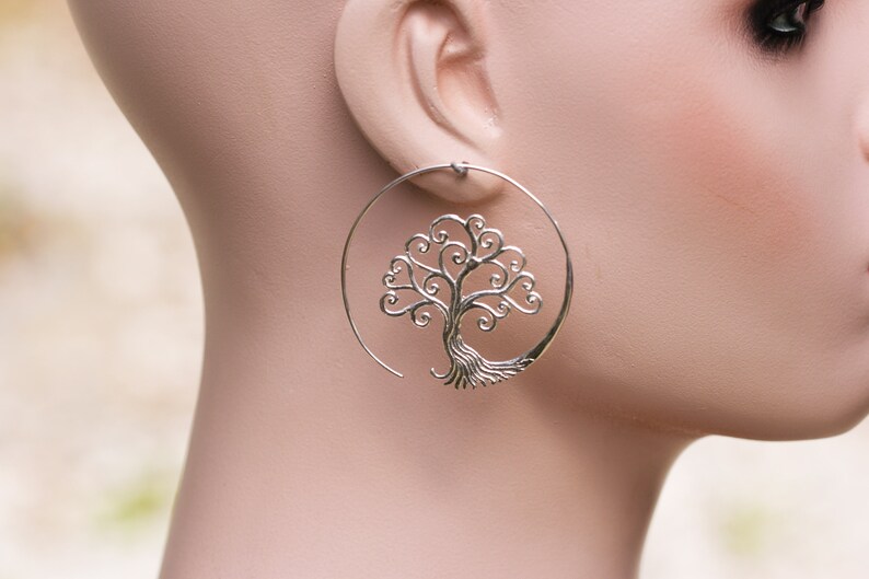 May include: Silver hoop earrings featuring a tree of life design. The tree is crafted from delicate swirls and branches.