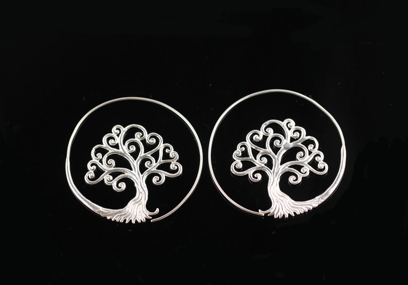 May include: A pair of silver hoop earrings featuring a tree of life design. The earrings are circular and have a detailed, intricate tree design with swirling branches.