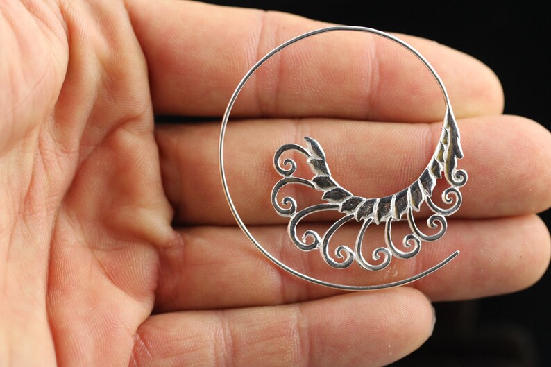 Op de afbeelding: Zilveren oorring met een decoratief, sierlijk ontwerp met een reeks swirls en bladeren.