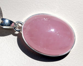 May include: A pendant necklace featuring a large, oval-shaped, polished rose quartz gemstone. The pink stone is set in a silver-colored metal bezel and attached to a silver chain. The pendant is a simple, elegant piece of jewelry.