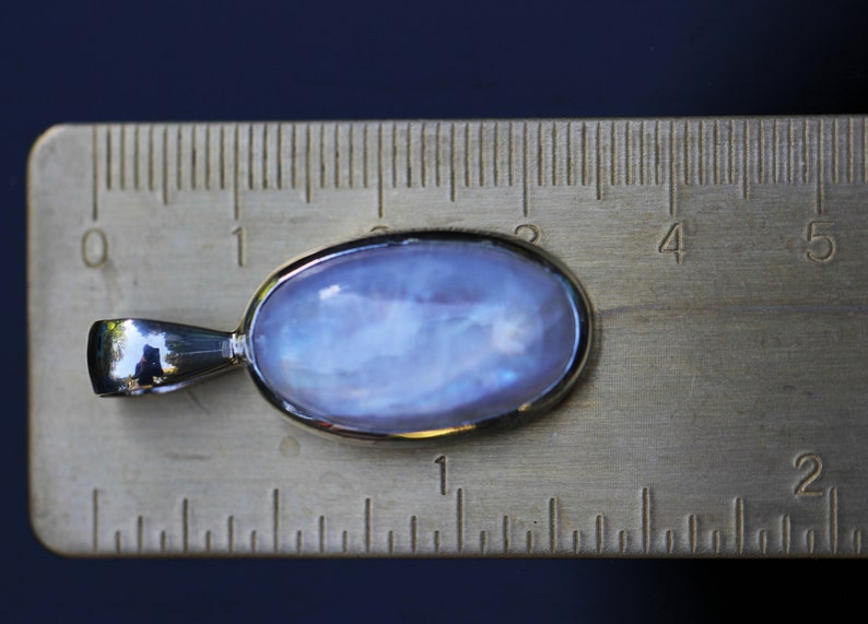 May include: A silver pendant with a large oval moonstone gemstone. The stone is a pale blue color with iridescent flashes of color. The pendant is about 1 inch long and is shown next to a ruler.